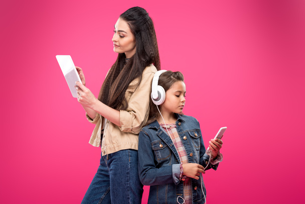 μητέρα και κόρη στην ακουστικά στέκεται πλάτη με πλάτη και ψηφιακές συσκευές που απομονώνονται σε ροζ  - Φωτογραφία, εικόνα