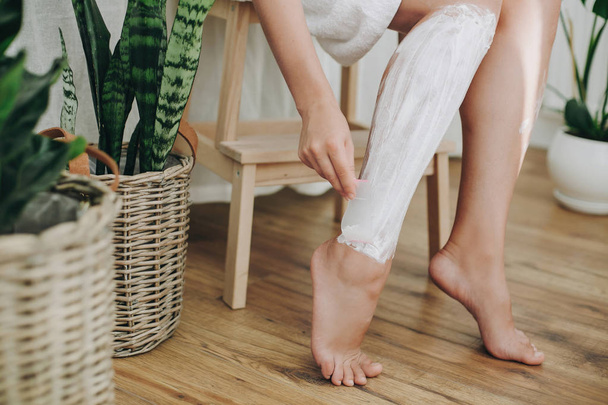 Hair Removal concept, depilation process. Young woman in white towel applying shaving cream on her legs and holding holding plastic razor in home bathroom with green plants. Skin care - Photo, Image