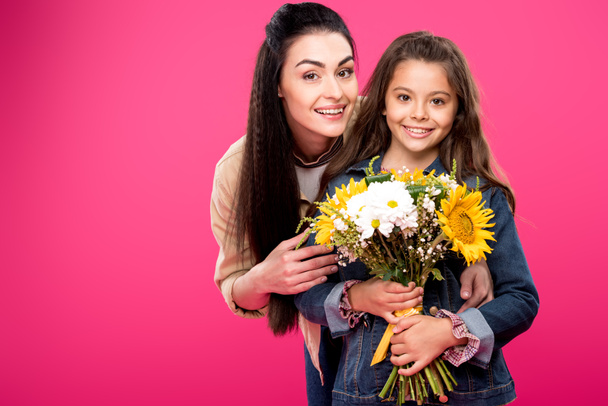 happy mother and daughter holding beautiful bouquet of flowers and smiling at camera isolated on pink  - Photo, image