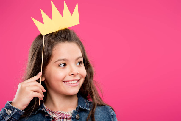 adorable happy child holding crown on party stick and looking away isolated on pink - Fotoğraf, Görsel