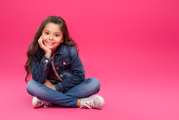 adorable happy child in denim clothes sitting with hand on chin and smiling at camera on pink - Φωτογραφία, εικόνα
