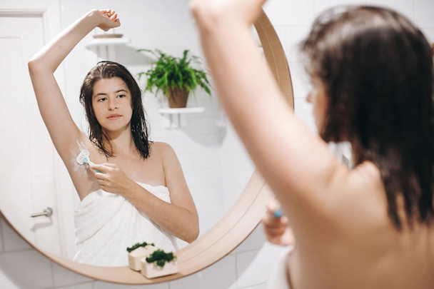 Haut- und Körperpflege. Haarentfernungskonzept. Frau nach der Duschrasur mit Rasiermesser. junge attraktive Frau in weißem Handtuch rasiert Achseln, Blick in den Spiegel in stilvollen Badezimmer. - Foto, Bild
