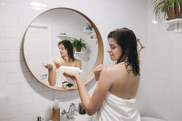 Φροντίδα του δέρματος και του σώματος. Νεαρή γυναίκα ευτυχισμένη στην λευκή πετσέτα, εφαρμογή ενυδατική κρέμα για ώμο στο μπάνιο, το είδωλό του στον καθρέφτη. Χέρι που κρατά το πλαστικό μπουκάλι με λοσιόν. Χαλαρωτικό σέξι γυναίκα - Φωτογραφία, εικόνα