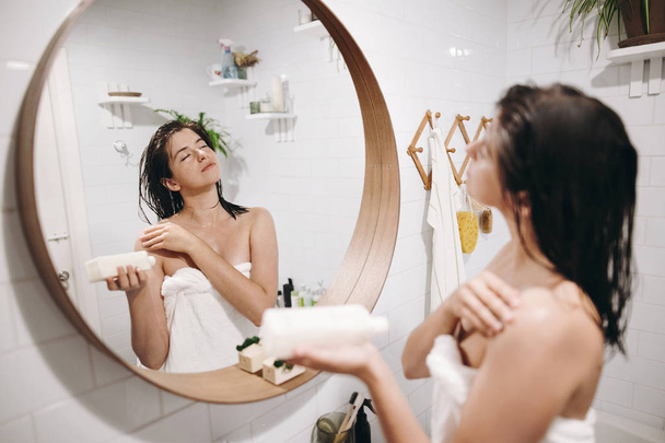 Cuidado de la piel y el cuerpo. Mujer sexy relajada en toalla blanca aplicando crema hidratante en el hombro en el baño, reflejo espejo. Mujer joven delgada disfrutando de spa y bienestar
 - Foto, imagen