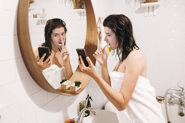 Daily morning routine. Young happy woman in white towel brushing teeth and looking at phone screen in stylish bathroom at round mirror. Slim sexy woman with natural skin and wet hair - Photo, Image