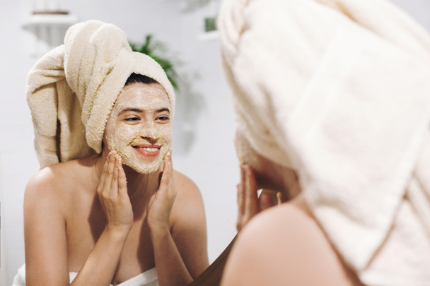 Концепция ухода за кожей. Молодая счастливая женщина в полотенце делает массаж лица с органическим скрабом лица и смотрит в зеркало в стильной ванной комнате. Девушка наносит крем, пилинг и чистку кожи
 - Фото, изображение