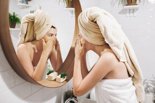 Νεαρή γυναίκα ευτυχισμένη σε πετσέτα εφαρμόζοντας scrub προσώπου βιολογικά και κοιτάζοντας στρογγυλό καθρέφτη στο κομψό μπάνιο. Κοπέλα κάνει μασάζ προσώπου, απολέπιση και καθαρισμός το δέρμα στο πρόσωπο. Φροντίδα του δέρματος και την υγιεινή - Φωτογραφία, εικόνα