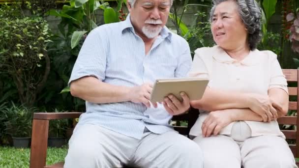 Senior couple sitting and using tablet together in home garden. Retired old Asian male and female, reading news in tablet, happy smile. Senior lifestyle concept.  - Felvétel, videó