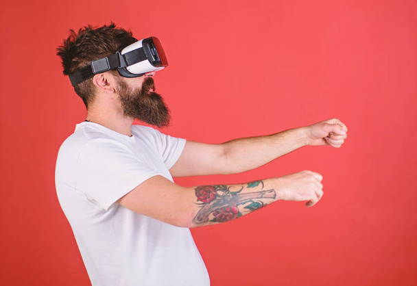 Hipster on confident face driving bike in virtual reality with modern digital gadget. Man with beard in VR glasses driving motorbike, red background. Virtual driving lessons concept - Foto, Imagen