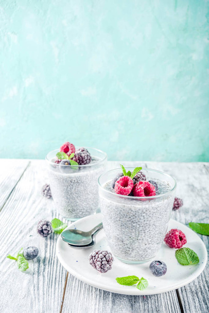 Summer healthy breakfast dessert in glass, overnight vegan chia pudding with non-dairy milk, frozen raspberry, blackberry and blueberry, on light blue and wooden background copy space - Zdjęcie, obraz