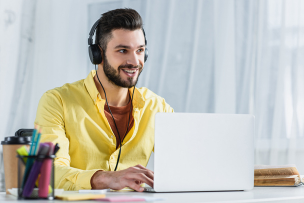 Smiling bearded businessman in headphones looking away at workplace - Fotó, kép