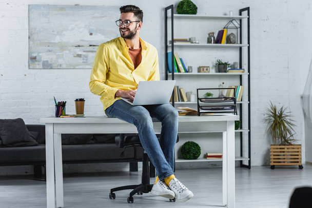 Bearded businessman in glasses typing, sitting on desk and looking away - Foto, Bild