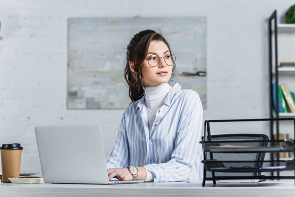 Attractive businesswoman in glasses using laptop and looking away  - Photo, Image