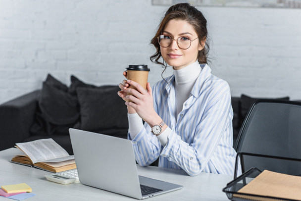 Attractive businesswoman holding paper cup, using laptop and looking at camera - Photo, Image