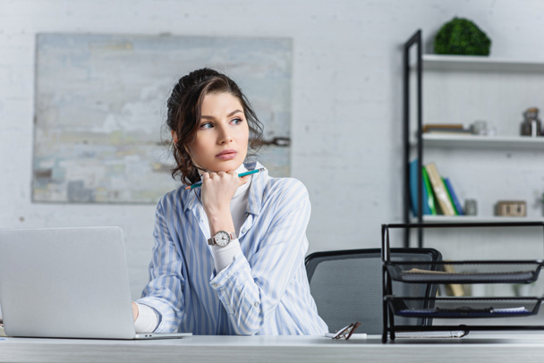 Beautiful businesswoman holding pencil, using laptop and looking away - Foto, afbeelding