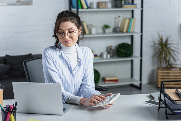 Veselá žena v brýlích, drží Kalkulačka u notebooku v moderní kanceláři  - Fotografie, Obrázek