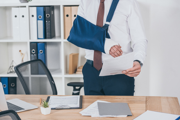 bijgesneden weergave van werknemer met gebroken arm in verband staande en papier te houden boven tafel in kantoor, compensatie concept - Foto, afbeelding