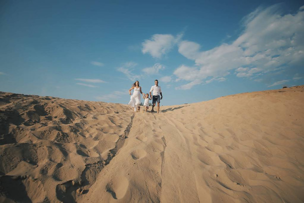 Cheerful caucasian family in white clothes with pretty daughter runs on desert sand dune. Pregnant woman - 写真・画像