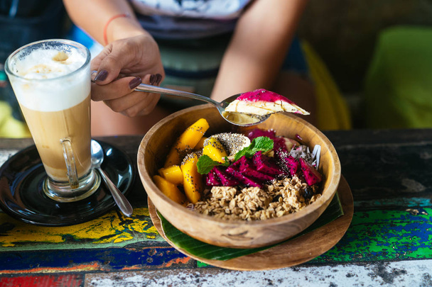 Womans hands with smoothie bowl made with mango, banana, granola, grated coconut, dragon fruit and mint with latte coffee - 写真・画像