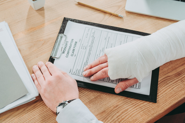 cropped view of worker with broken arm in gypsum holding hands on compensation form in office, compensation concept - Фото, изображение