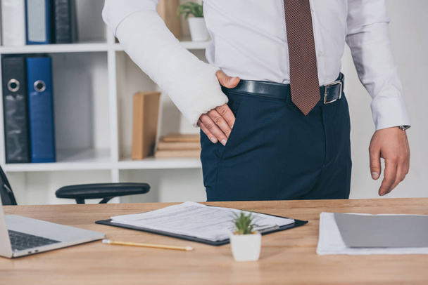 cropped view of worker with broken arm holding hand in pocket and standing at table in office, compensation concept - 写真・画像