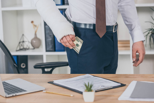 cropped view of worker with broken arm geting money out of pocket and standing at table in office, compensation concept - Photo, Image
