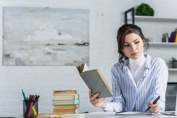upset woman studying with book while holding pen in hand in modern office  - Foto, Imagen