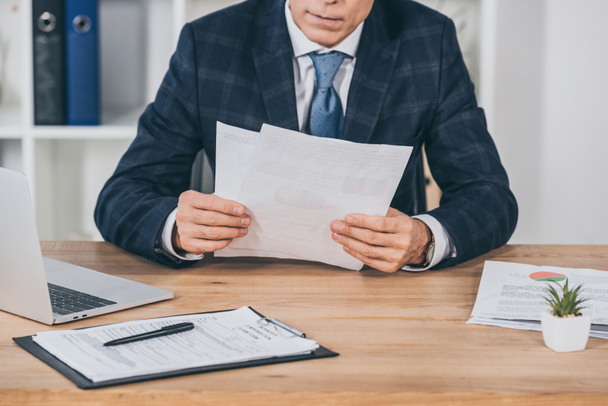 cropped view of businessman in blue jacket sitting at table and reading documents in office, compensation concept - Foto, imagen
