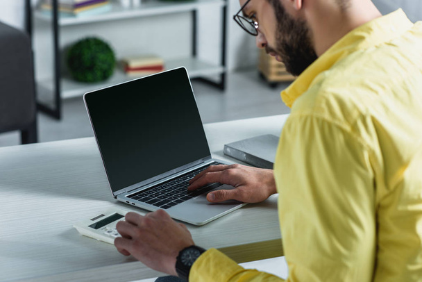 bearded man looking at calculator near laptop with blank screen in modern office - Fotó, kép