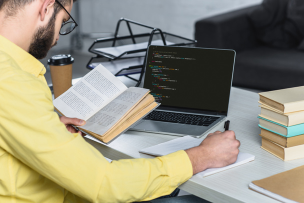 homem barbudo estudando com livro perto de laptop com codificação de computador na tela no escritório moderno
 - Foto, Imagem