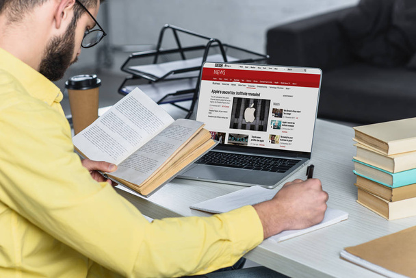 бородатый человек учится с книгой возле ноутбука с bbc сайта на экране в современном офисе
 - Фото, изображение