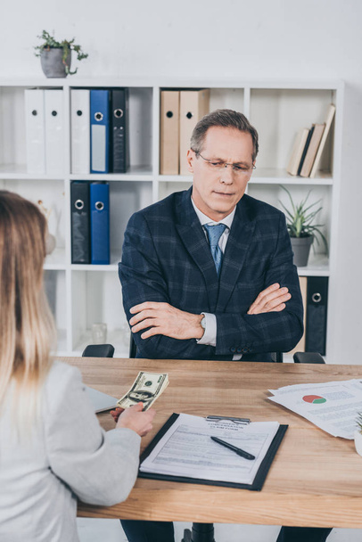 бизнесмен среднего возраста, сидящий за столом с документами и глядя на деньги, предоставляемые женщиной в офисе, концепция компенсации
 - Фото, изображение