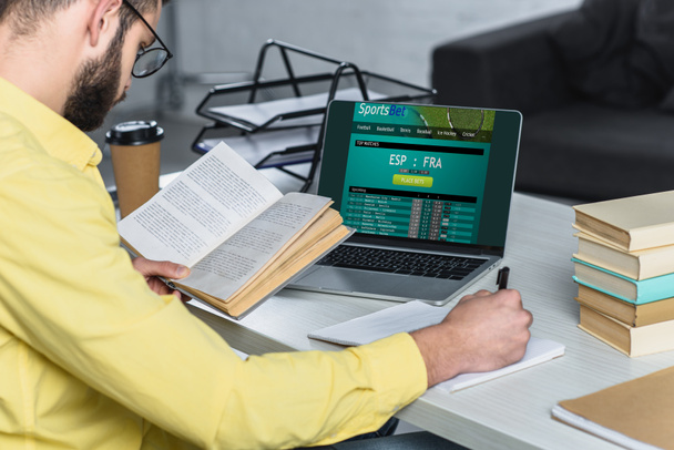 uomo barbuto studiare con libro vicino laptop con sito sportsbet sullo schermo in ufficio moderno
 - Foto, immagini