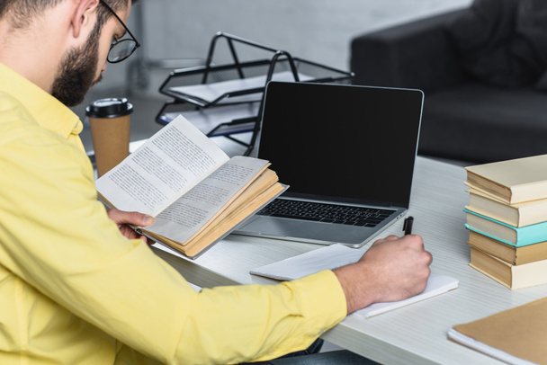 человек учится с книгой возле ноутбука с чистым экраном в современном офисе
 - Фото, изображение