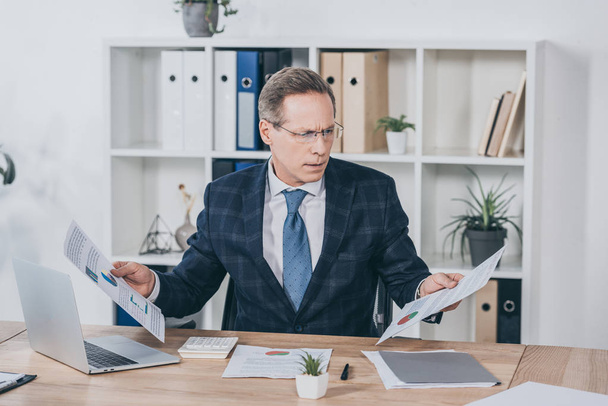middle aged businessman sitting at table, holding documents and reading in office, compensation concept - Photo, Image