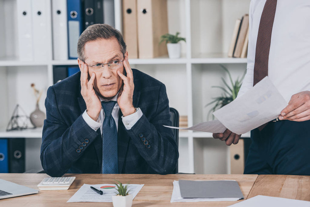 middle aged dissatisfied businessman sitting at table wile worker standing near  with documents in office, compensation concept - Фото, изображение