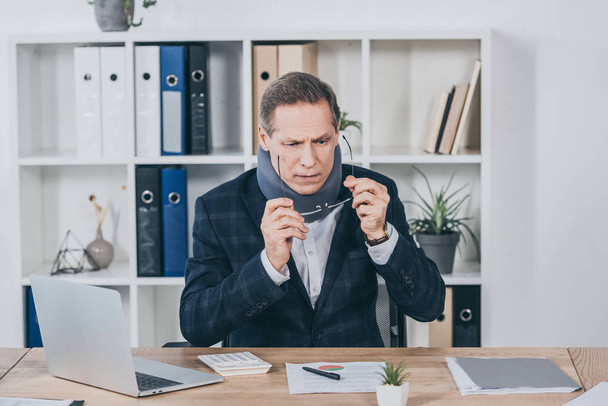 businessman in neck brace sitting at table wiht documents, puting on glasses in office, compensation concept - Photo, Image