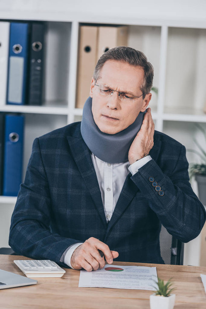 businessman in neck brace sitting at table, feeling pain and holding neck in office, compensation concept - Valokuva, kuva