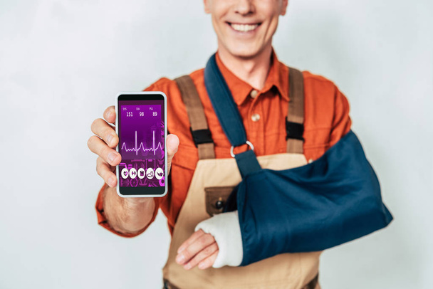 обрезанный вид ремонтника со сломанной рукой и бинтом, показывающий смартфон на белом фоне
 - Фото, изображение