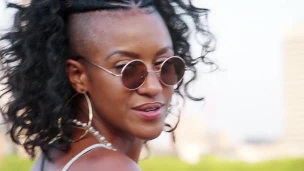 Trendy young black woman in camisole and sunglasses walking over bridge, looking back and laughing to camera, close up, follow shot - Filmati, video