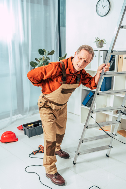 middle aged repairman standing and holding on ladder and back in office - Photo, Image
