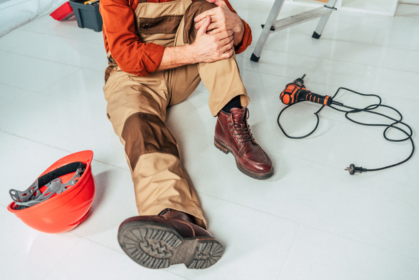 cropped view of repairman sitting on floor and holding injured knee in office - 写真・画像