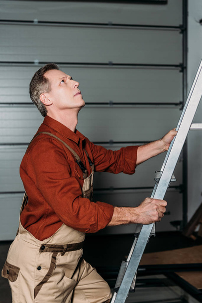 reparateur in oranje uniforme klimmen op de ladder in garage - Foto, afbeelding