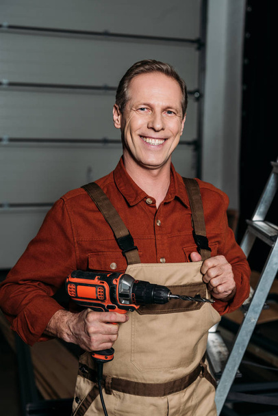 repairman standing with screwdriver near ladder and smiling in garage - Foto, afbeelding
