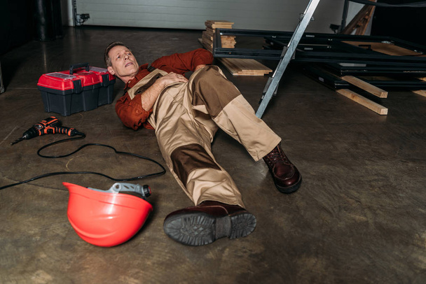 επισκευαστής, ξαπλωμένο στο πάτωμα με τη στολή κοντά Σκάλα και κρατώντας στα πλευρά στο γκαράζ - Φωτογραφία, εικόνα