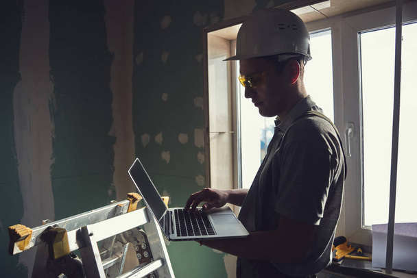 Room repair. Builder in a helmet and glasses stands in profile with a laptop in hand on the background of the construction site - Foto, immagini