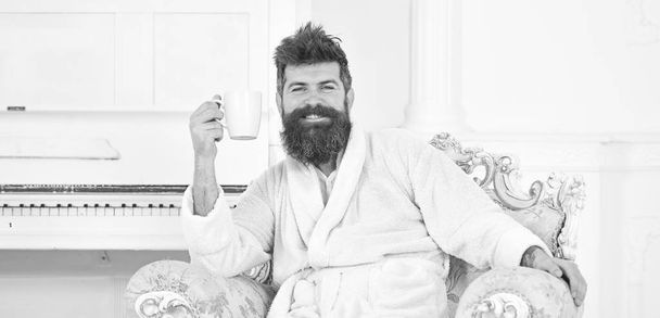 Cheerful guy in white bathrobe enjoying morning coffee. Bearded man sitting in beautiful antique armchair. Energetic morning at home - Zdjęcie, obraz