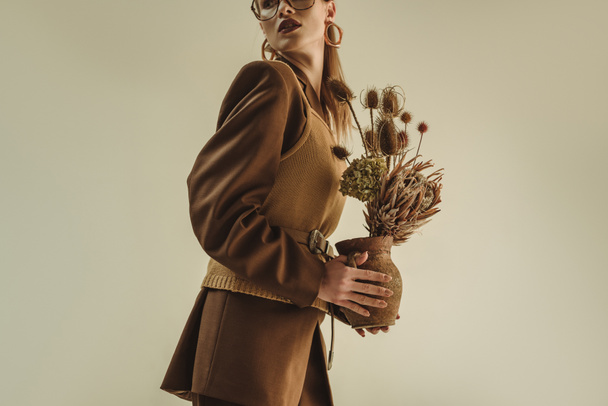 hermoso modelo en estilo retro sosteniendo jarra de arcilla con ramo de flores secas aisladas en beige
 - Foto, Imagen