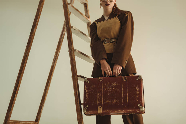 vista ritagliata della ragazza in posa con borsa da viaggio vintage vicino alla scala isolata sul beige
  - Foto, immagini
