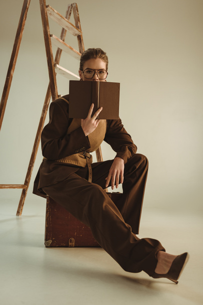 attractive model holding book and sitting near ladder on beige - Photo, Image